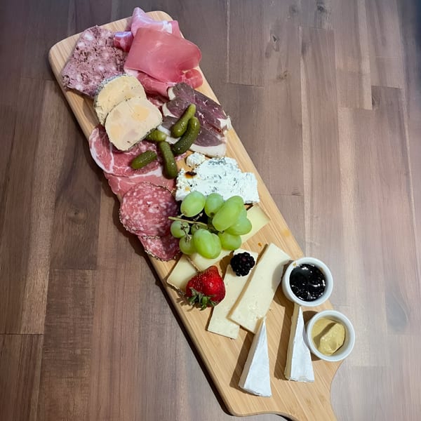 A long wooden charcuterie board filled with various meats, cheeses, grapes, berries, cornichons, and sauces.