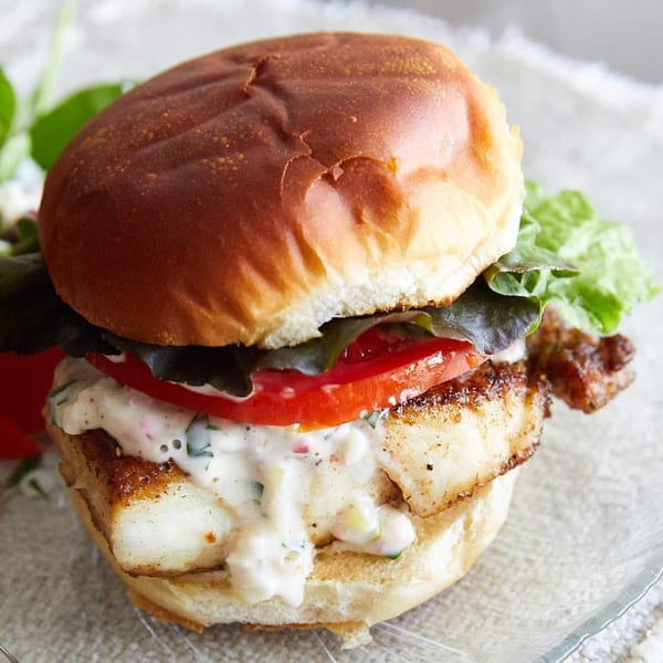 Close-up of a fish sandwich with lettuce, tomato, and creamy sauce on a brioche bun.