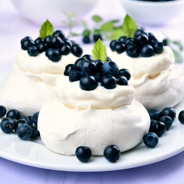 Three meringue desserts topped with whipped cream, fresh blueberries, and mint leaves on a white plate.