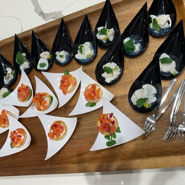 A wooden tray holds black teardrop spoons with ceviche and white triangular plates with bruschetta.