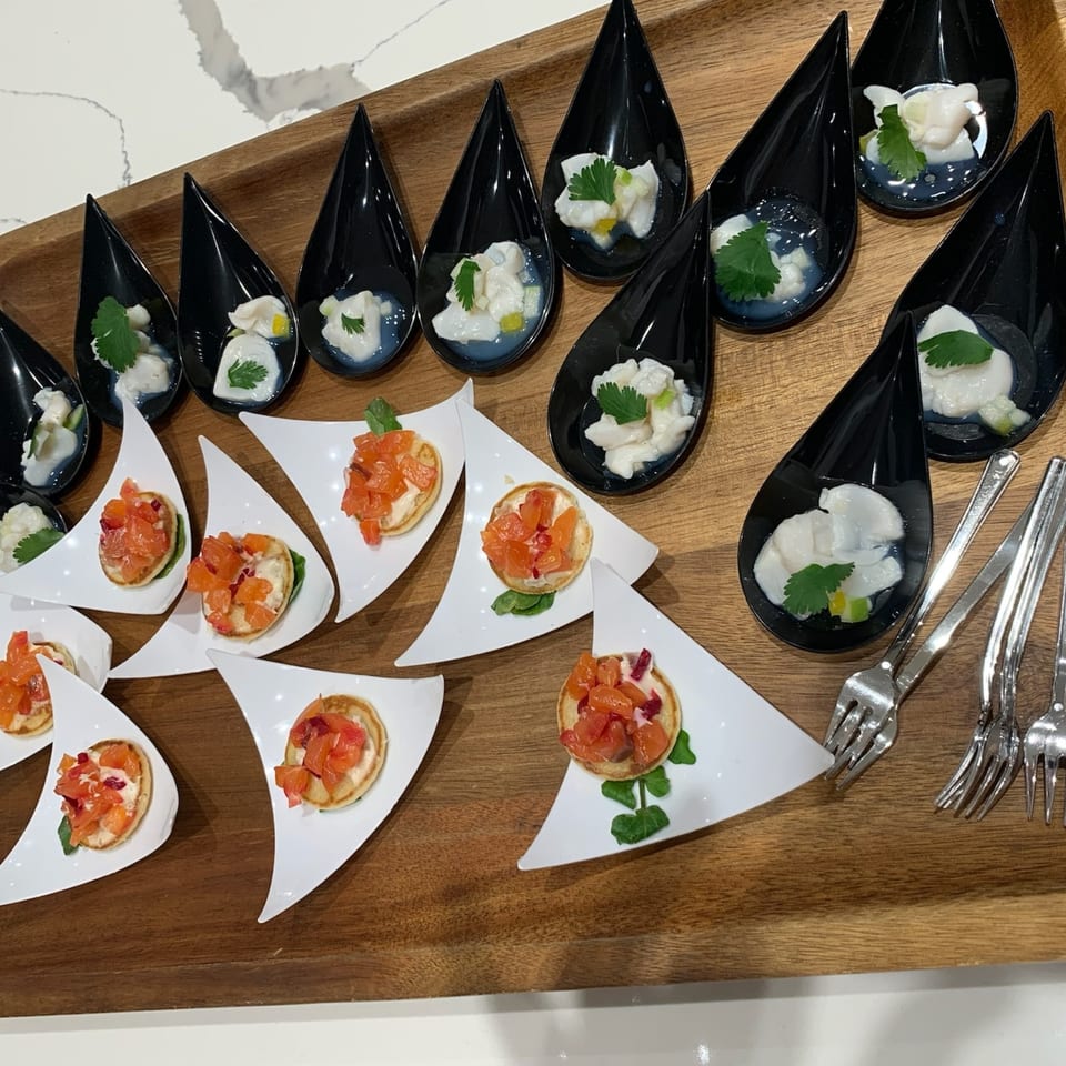 A wooden tray holds black teardrop spoons with ceviche and white triangular plates with bruschetta.