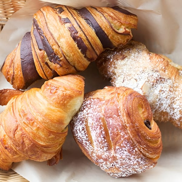 Assortment of golden-brown croissants, including pain au chocolat and almond, in a paper-lined basket.